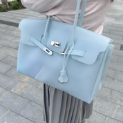 A light blue structured handbag with silver-tone hardware, carried on a person wearing a pale pink top and gray pleated skirt, photographed outdoors on a paved walkway.