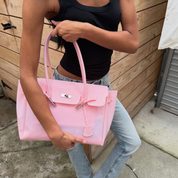 A woman wearing a black tank top and light blue jeans holds a large light pink structured handbag with silver-tone hardware in front of a wooden wall.
