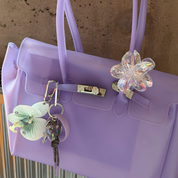 A lavender translucent jelly handbag with a gold turn-lock clasp, decorated with a light green orchid keychain charm and a clear iridescent flower charm, set against a textured concrete wall.
