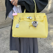 A person holds a large structured yellow handbag outdoors. The bag has a flap top, top handles, a small pink pouch charm, a white lock detail, and two hanging charms: a small white house keychain and a gray plush character.