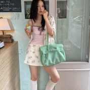 A woman stands indoors holding a mint green structured handbag with silver hardware. She wears a pale pink top, a cream floral skirt, and white knee-high socks, with a lamp and wall visible in the background.